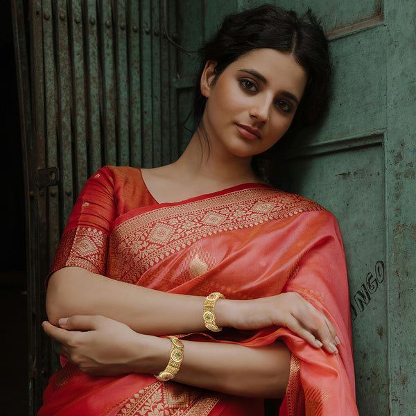 Smiling model wearing a Punjabi kangan bangle pair in gold-tone with red and green stones.
